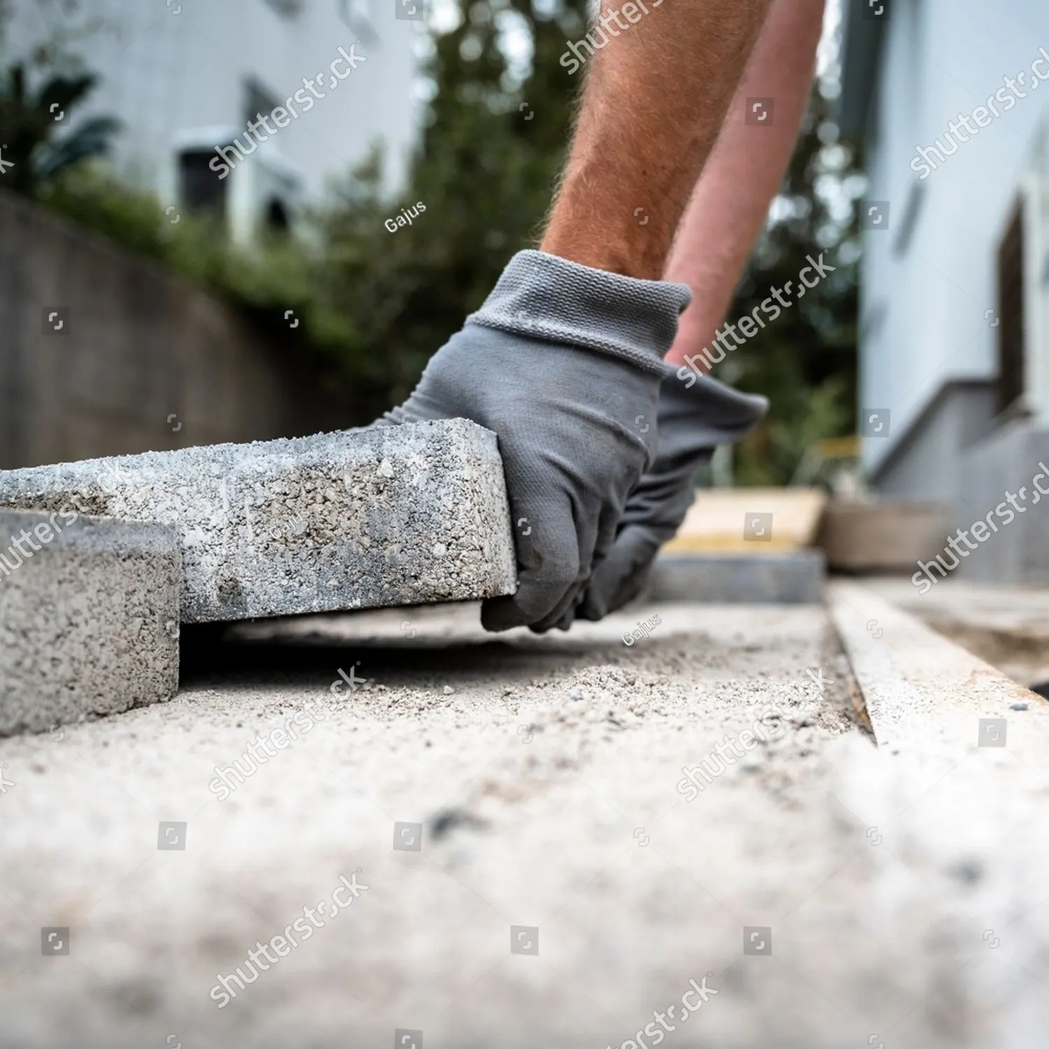 Dłonie w rękawicach roboczych układają płytę betonową na podsypce